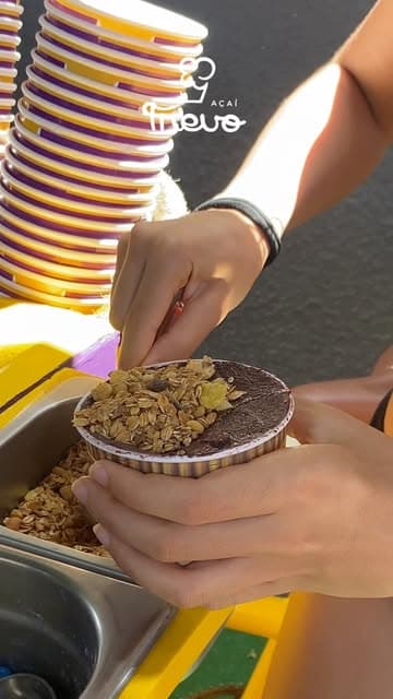 Mãos montando um copo de açaí Trevo com granola e complementos, com torre de copos ao lado